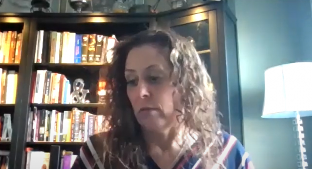 Colour screen capture of a woman looking down with a thoughtful expression. Head and shoulders are visible, with shelves of books in the background.