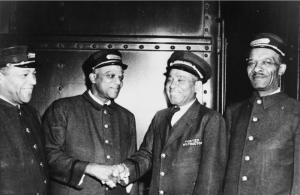 Black and white archival photograph of four Black men standing in a row. The two men in the centre are shaking hands and all four men are smiling.
