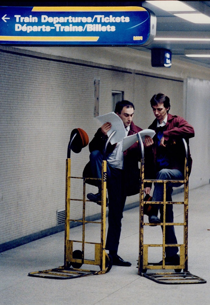 Colour archival photograph of two men in railway uniforms leaning on two yellow baggage carts while examining papers. There is a red cap hanging from one of the baggage carts.