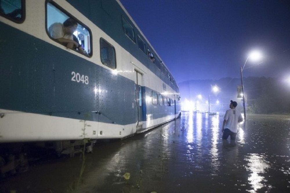 Colour photograph of a partially submerged GO Train in brown water. There is a passenger leaning out of an open window on the train. A man is standing in water up to his knees next to the train and looking up at the train.