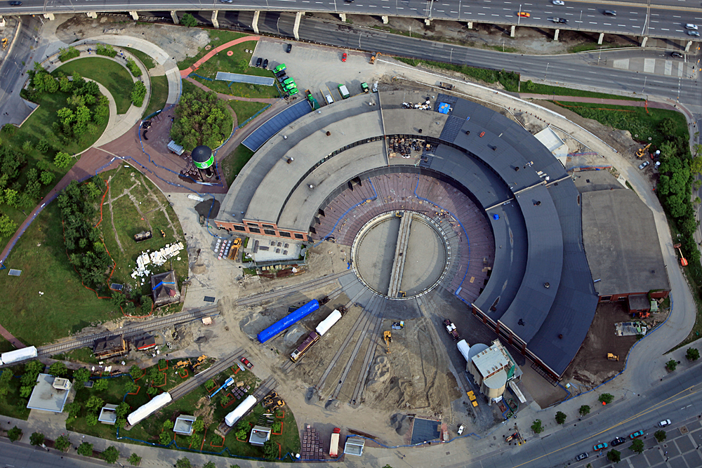 Modern colour aerial photograph showing a roundhouse building and railway tracks. There is a highway at the top of the photo and many rail cars and small buildings around the roundhouse. There is a park to the left of the roundhouse.