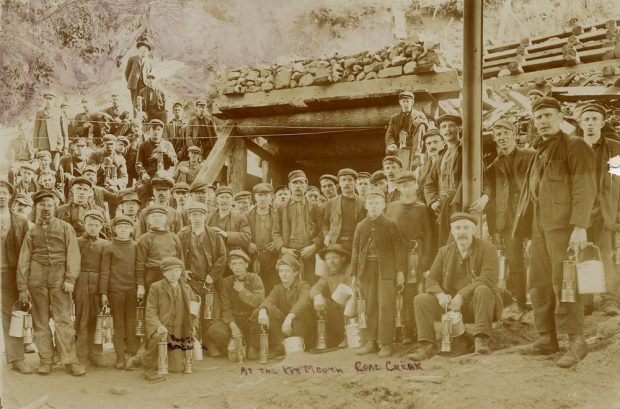 Group of miners holding mine lamps and lunch buckets in front of a mine entrance.