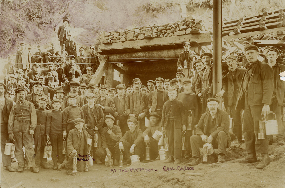 Group of miners holding mine lamps and lunch buckets in front of a mine entrance.