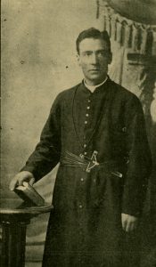 An Oblate missionary standing in his robes with a bible in his right hand and leaning on a table. with a heavy velvet and tassel curtain as a backdrop.