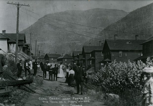 Men and women walking along a street. Wood sided houses are on both sides