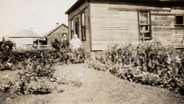 An elderly man and woman standing by the front door of their house; a large garden surrounds the house.