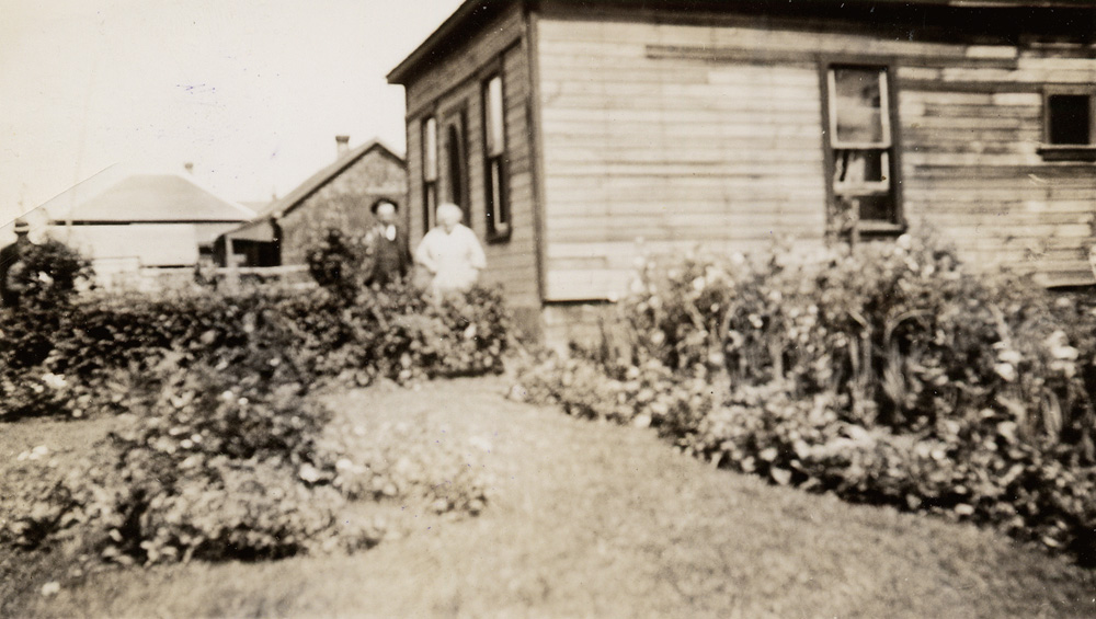 An elderly man and woman standing by the front door of their house; a large garden surrounds the house.
