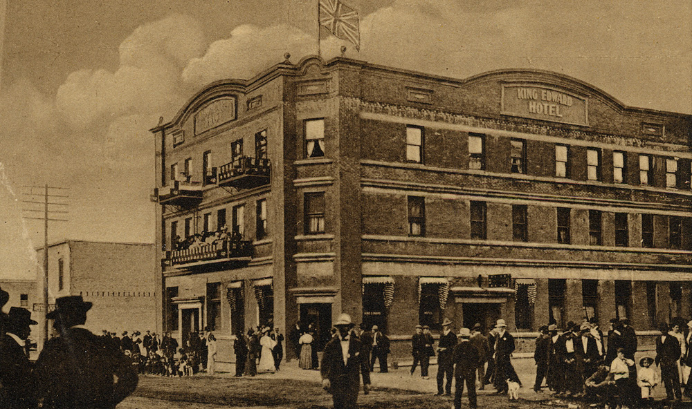 The three-story King Edward Hotel with a large crowd of men and women standing on the hotel balcony and in front of the hotel.