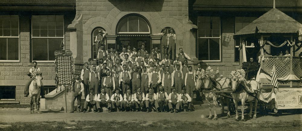 Five rows of men standing or kneeling in front of a building. Man on a horse is on the left, a parade float on the right.