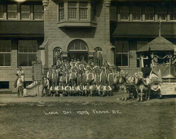 Five rows of men standing or kneeling in front of a building. Man on a horse is on the left, a parade float on the right.