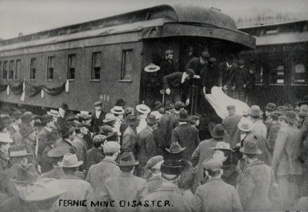 Large crowd of men and women in front of a railway passenger car. A dead body covered in a sheet is being unloaded from a train car.