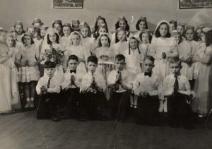 Quatre rangées d'enfants. Les filles sont en robes blanches et voiles. Les garçons sont en chemises blanches avec des cravates et des pantalons foncés.