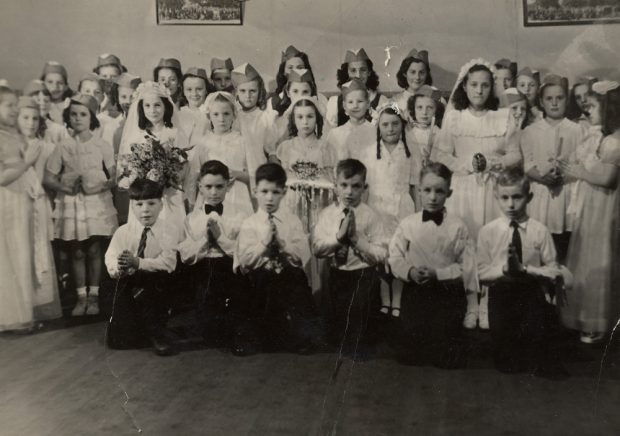 Four rows of children. Girls are in white dresses and veils. Boys are in white shirts with ties and dark pants.