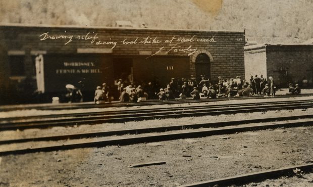 Three rows of men in front of a brick building; front row of men are kneeling.