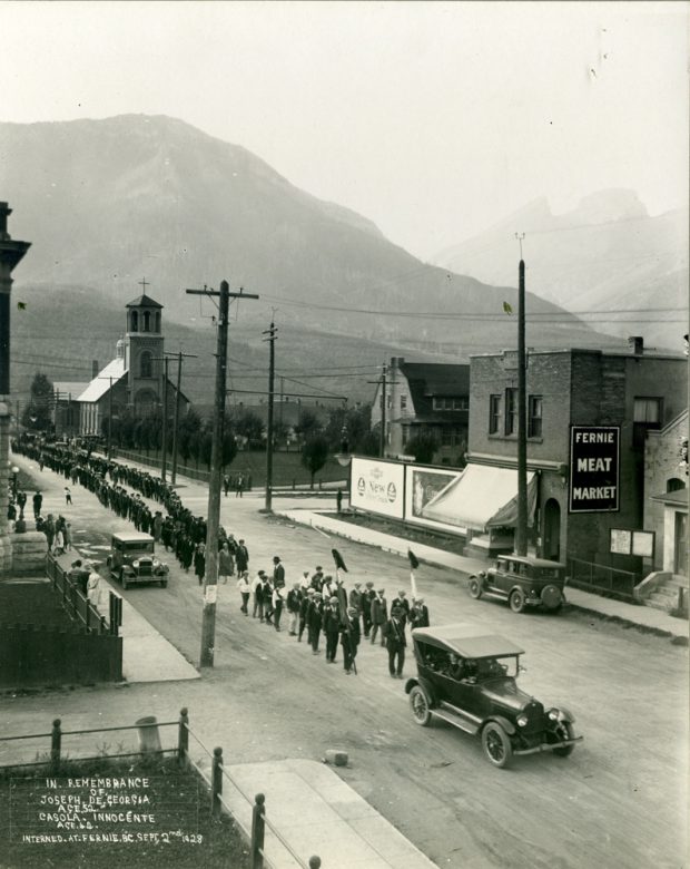 A car is leading a large funeral procession. Men are walking in two's behind the car.