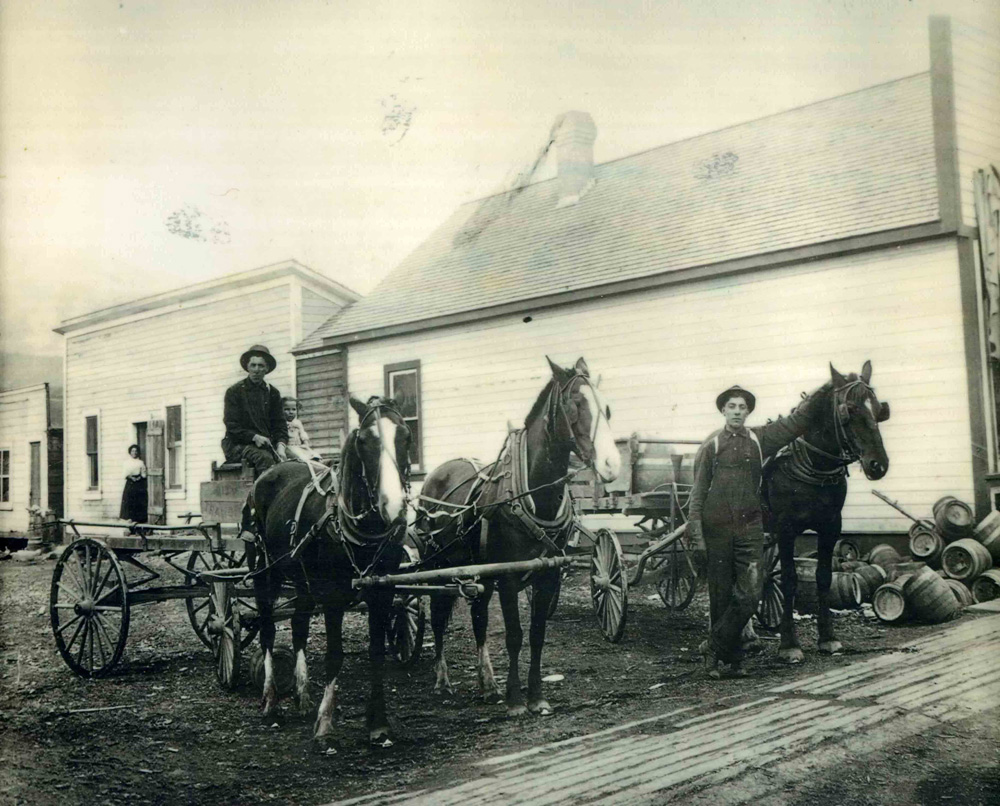 A store in the background with a wagon with horses in the front.