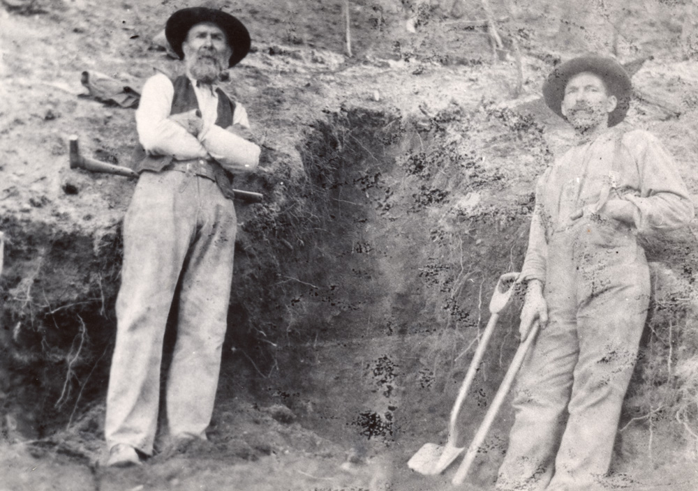 Two men stand in front of a hole in a hill. The man on the right has a pick and a shovel.