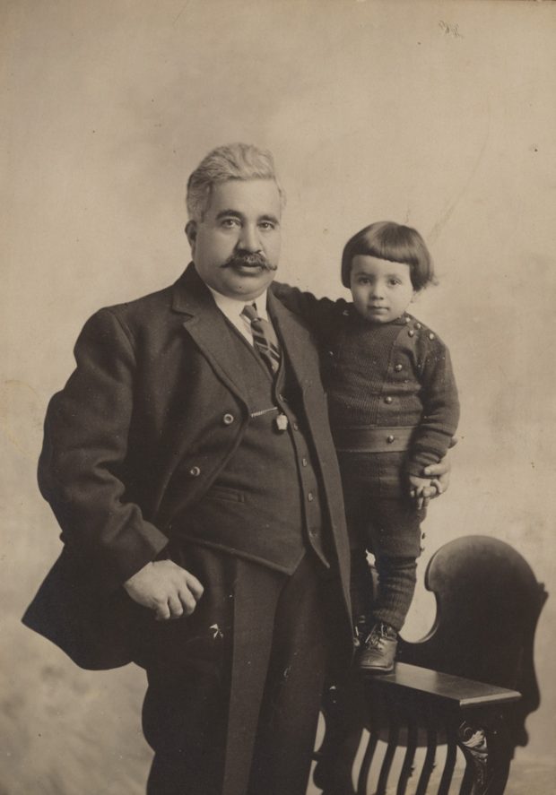 A man with a moustache standing next to a young boy on a chair.