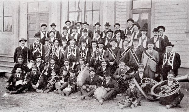 Five rows of men; front two rows of men are holding instruments and are sitting. Three rows of men in regalia stand behind them.