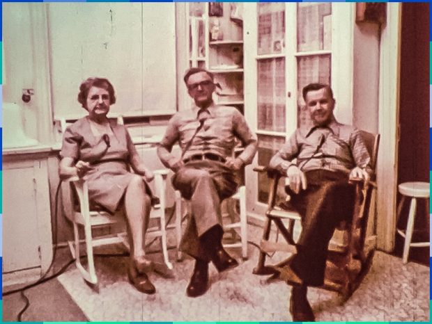 A woman and two older men are sitting on chairs or rocking chairs in a kitchen. They are looking at the camera.