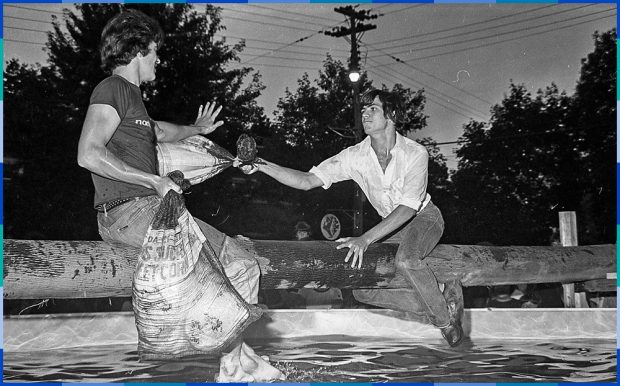 Two young men are sitting on a log hanging over a pool. Each of them is holding a bag of corn, which they are using to hit each other. Curious onlookers are gathering around the pool to watch.