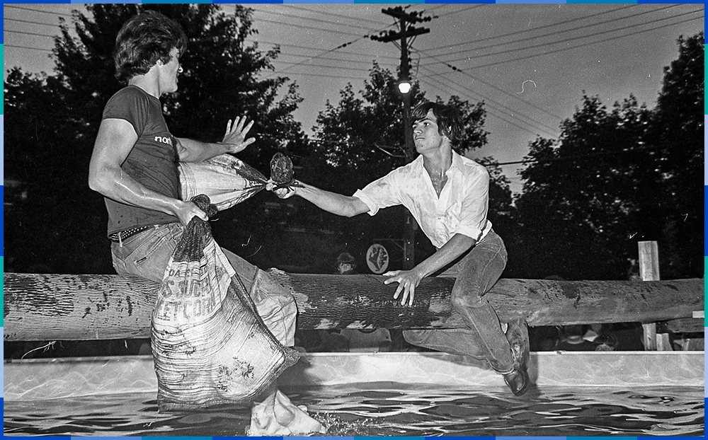 Two young men are sitting on a log hanging over a pool. Each of them is holding a bag of corn, which they are using to hit each other. Curious onlookers are gathering around the pool to watch.