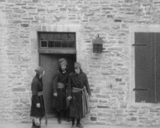 Three patriots wearing toques and long coats fastened with arrow sashes are at the door of a stone building.
