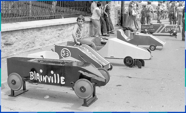 A racing car (soapbox) has been placed on blocks. A child is sitting on the second soapbox from the left. A crowd can be seen in the background on the right.