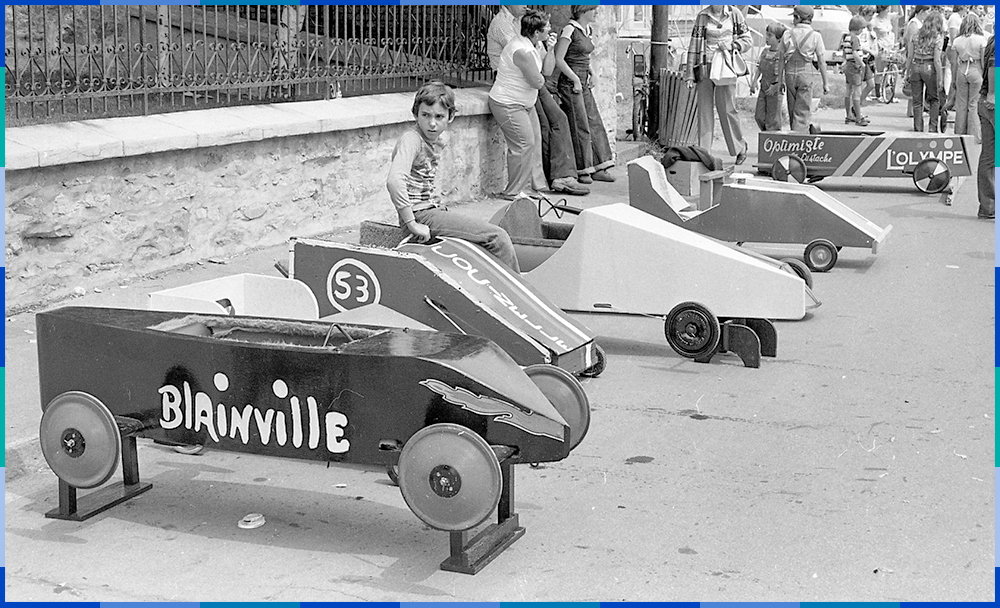A racing car (soapbox) has been placed on blocks. A child is sitting on the second soapbox from the left. A crowd can be seen in the background on the right.