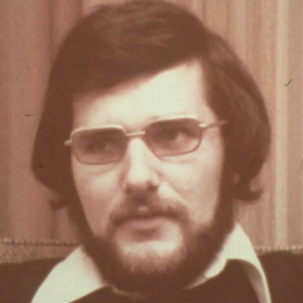A photographic portrait of a man with a beard and medium-length hair, wearing glasses. A shirt collar can be seen above his sweater.