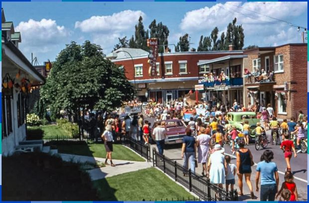 A colour photograph of a crowd of people of all ages parading down a shopping street. A few children are on bicycles, while the others are on foot. There are around ten people watching the street from two second-storey balconies over two shops.