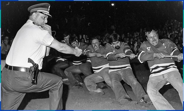 Five men wearing identical sweaters are pulling on a thick rope. Their bodies are bent backwards by the effort. In the foreground, a police officer with a whistle in his mouth referees the activity, and in the background is a crowd of spectators.
