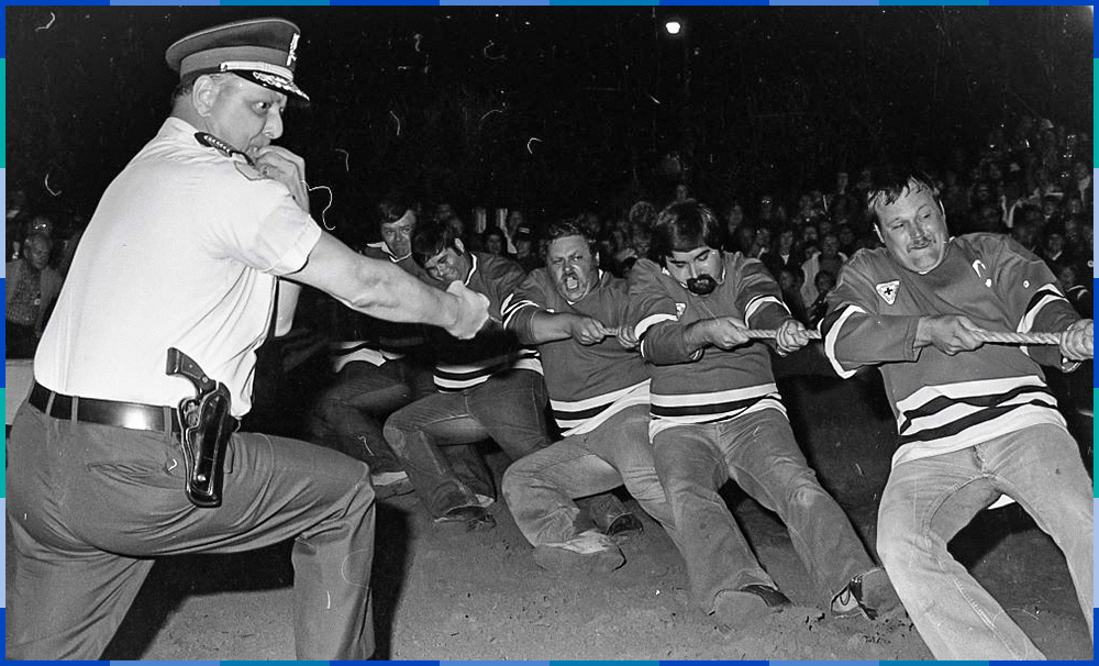 Five men wearing identical sweaters are pulling on a thick rope. Their bodies are bent backwards by the effort. In the foreground, a police officer with a whistle in his mouth referees the activity, and in the background is a crowd of spectators.