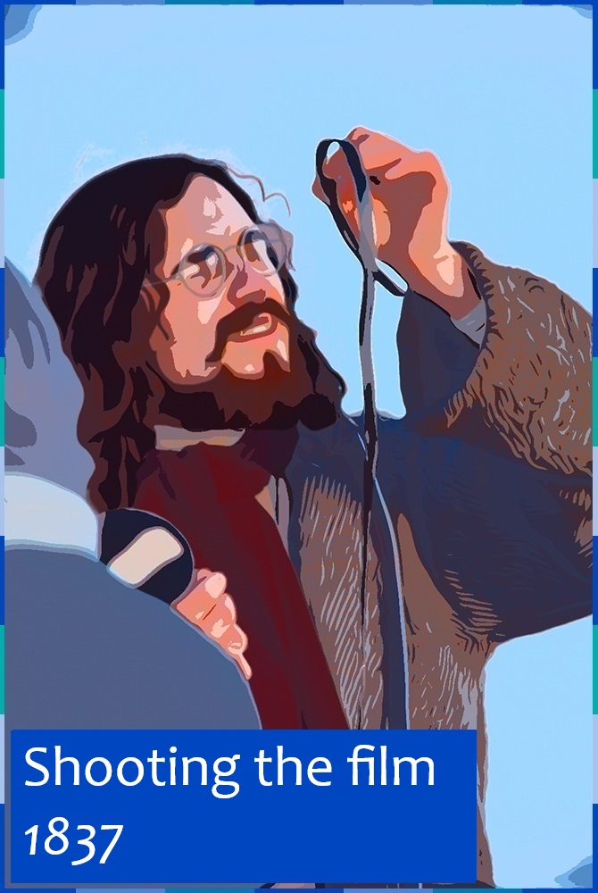 A photograph, edited to look like a comic strip, of a man with a beard and long brown hair. He is wearing a coat, a burgundy scarf, and small round glasses. He is examining a roll of film. On the lower left corner are the words: “Shooting the film [1837]”.