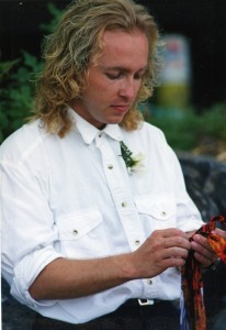 Man in white western style shirt with a white rose corsage. Holds a piece of bright coloured fabric.