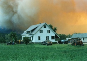 White house with vehicles parked close by, people near their vehicles. Sky is a yellow-orange colour. Fire burns on hillside.