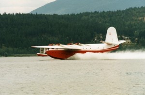 Red and white float plane lands in water. Hillside in background.