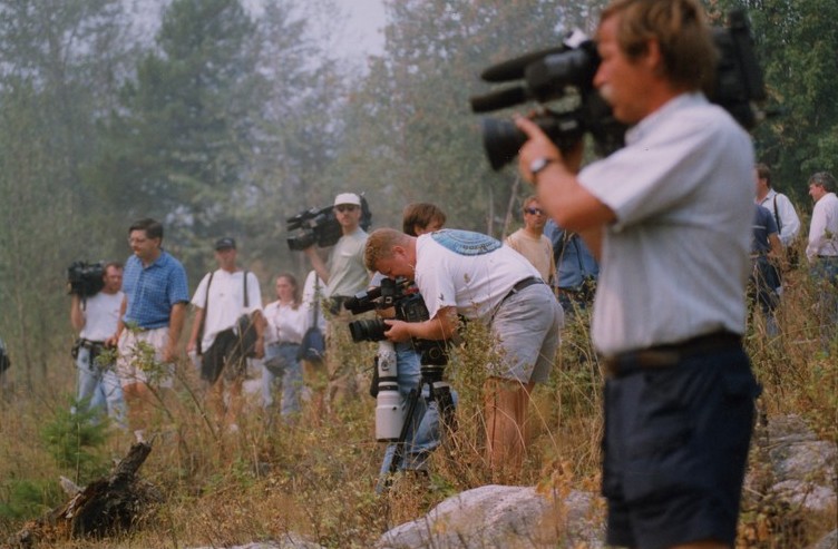 13 reporters in brush, some with large cameras over their shoulders.