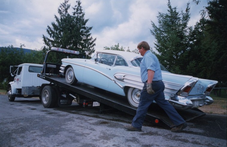 Blue and white collector’s car loading on a flatbed hitched to a tow truck. Man walks in front of car.