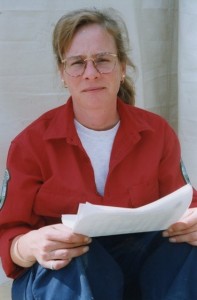 Seated blonde woman with glasses looks at the camera. She is holding a paper.
