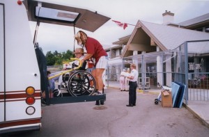 An attendant unloads a person in awheelchair from a bus. Two other women walk towards a building's entrance. A person in a uniform surveys the scene.