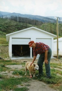 Man pets his dog in front of a white garage. Dog has a bandage on its paw.