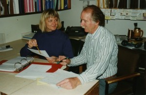 Man and woman sit at a desk consulting papers in an office.