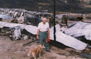 Man and dog stand in front of rubble and twisted metal.