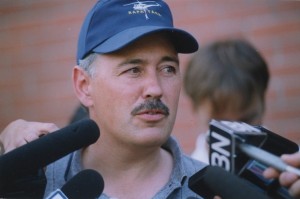 Man in with a mustache, wearing a grey golf shirt and a blue Rapattack baseball cap stands before many microphones.
