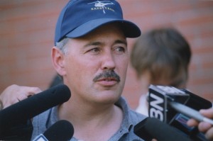 Portrait of a mustached man in a blue baseball cap speaking into microphones.