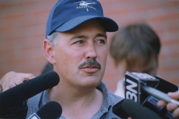Portrait of a mustached man in a blue baseball cap speaking into microphones.