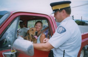 Woman with her dog in her lap sits in a red pick-up truck. RCMP officer talking to her. Another passenger in the truck sits with a dog in his lap.