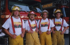 Four firefighters stand in front of their fire trucks.