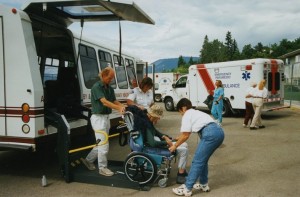 Two workers and a uniformed person assist a man in a wheelchair. Others are walking to and from the front of the bus. Two ambulances in background.